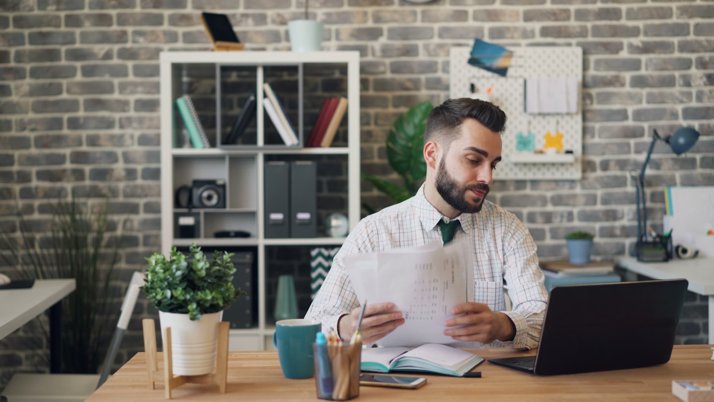 Man reviews documents at a desk with a laptop and plant.